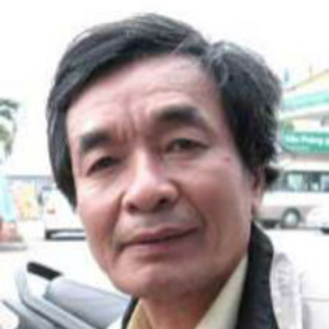 A middle-aged man with short, dark hair and wearing a light-colored jacket stands outdoors, with cars and buildings visible in the blurred background.