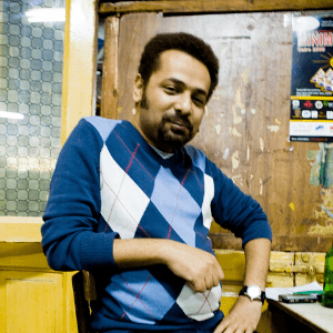 A man with short hair and a beard, wearing a blue argyle sweater, sits indoors at a table. The background includes wooden panels, a glass window, and a wall with posters.
