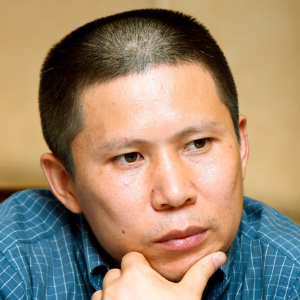 A person with closely cropped hair and a blue checked shirt rests their chin on their hand, looking thoughtful and serious against a neutral background.