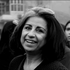 A woman with shoulder-length dark hair and streaks of gray smiles while standing outdoors in a crowd. She is wearing a knit sweater and has sunglasses resting on her head. The image is in black and white.