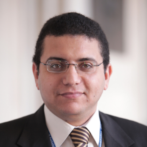 A man with short dark hair and glasses, wearing a dark suit, white shirt, and striped tie, is facing the camera and smiling slightly. The background is softly blurred.