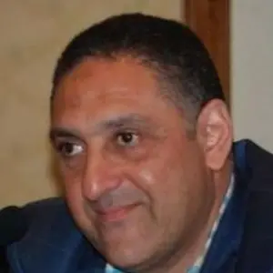 A middle-aged man with short dark hair, wearing a blue jacket and checkered shirt, is looking slightly to the side and smiling softly. The background is out of focus.