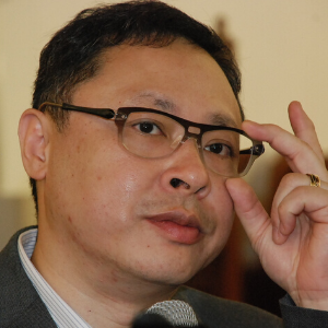 A man with short dark hair and glasses adjusts his frames with his right hand. He is wearing a suit jacket and looking slightly to the side.
