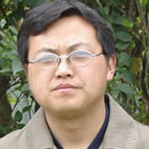 A man with short dark hair and glasses is standing outdoors in front of green foliage, wearing a beige jacket over a dark shirt.