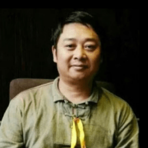 A person with short dark hair, wearing a green shirt with a yellow ribbon tied at the collar, is sitting and smiling slightly against a dark background.