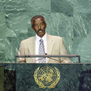 A man in a tan suit and patterned tie stands at a podium with the United Nations emblem, speaking in front of a green marble background.