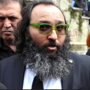 A man with a thick, dark beard and long hair, wearing bright green glasses, a suit, and a black tie, stands outdoors. Another man is visible in the background, partially out of focus.