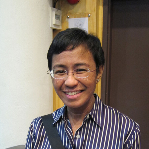 A person with short black hair and glasses smiles while wearing a blue and white striped shirt, standing indoors near a wall and a brown door.