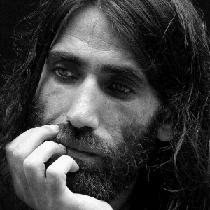 A man with long hair and a beard rests his face on his hand, looking thoughtful and introspective. The image is in black and white.