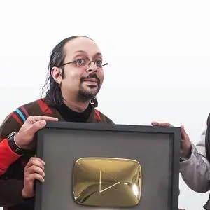 A man with glasses and shoulder-length hair, wearing a striped sweater, holds a framed YouTube Gold Play Button award with the help of two other people, against a plain white background.