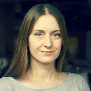 A woman with long straight brown hair and light eyes is looking at the camera, wearing a light grey top, with a subtle smile and a neutral, softly lit background.