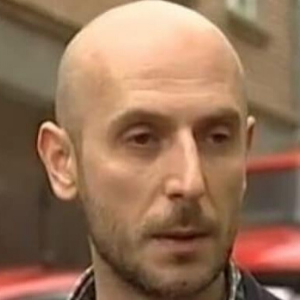 A bald man with light skin and a trimmed beard is outdoors, standing in front of a blurred background with a red vehicle and a building visible.