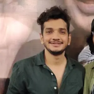 A man with short, curly hair and a trimmed beard is smiling while wearing a dark green shirt with an open collar, standing in front of a blurred background.