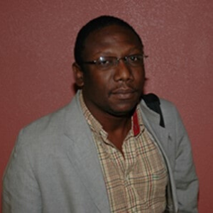 A man with short hair and glasses wearing a light gray blazer over a beige plaid shirt stands in front of a reddish-brown wall, looking at the camera.