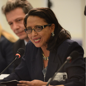 A woman with glasses, wearing a dark blazer and necklace, speaks into a microphone at a conference table. Other people are seated beside her, and she holds papers in her hands.