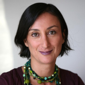 A woman with short black hair and purple lipstick smiles slightly at the camera. She is wearing a maroon top and multiple beaded necklaces. The background is plain and softly lit.