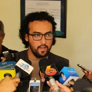 A man with curly hair and glasses speaks to the media while several microphones are held in front of him. A blurred sign is visible on the wall in the background.