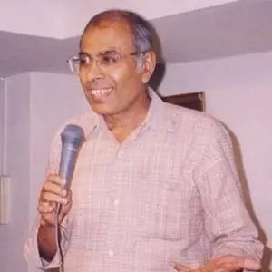 An older man with short gray hair and glasses is smiling while holding a microphone, wearing a light-colored shirt, and standing indoors.