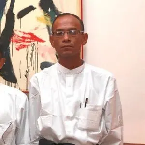 A man wearing glasses and a traditional white shirt stands indoors in front of an abstract painting. He has short hair and pens in his shirt pocket.
