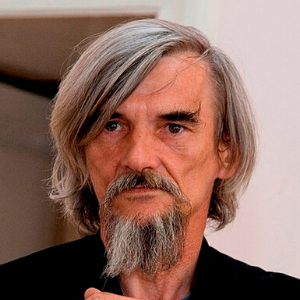 A middle-aged man with long gray hair and a full gray beard stands indoors, wearing a dark jacket. He has a serious expression on his face.