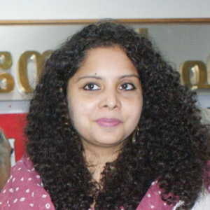 A woman with curly black hair wearing a maroon and white patterned top is looking slightly to the side. She is indoors, and a partial gold and white sign is visible in the background.