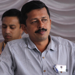 A man with short hair and a mustache, wearing a light gray button-up shirt with pens and a smartphone in the pocket, sits indoors. Another man is blurred in the background. The setting appears professional.