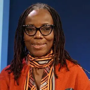 A woman with long braided hair and glasses smiles at the camera. She is wearing an orange jacket and a striped scarf, with a dark blue background behind her.