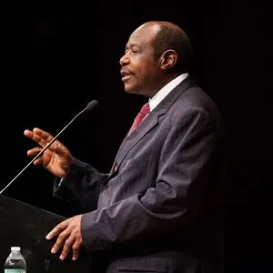 A man in a suit and tie speaks at a podium with a microphone, gesturing with one hand. A water bottle is visible on the podium against a dark background.
