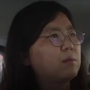 A person with long dark hair and round glasses looks slightly upward with a neutral expression while sitting indoors.