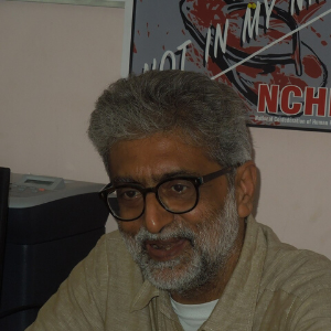 A middle-aged man with gray hair, a beard, and glasses smiles while sitting indoors. There is a poster and an office printer visible in the background.