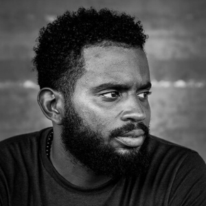 Black and white portrait of a man with short curly hair and a beard, gazing to his right with a serious expression. The background is plain and slightly blurred.