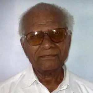 An elderly man with gray hair wearing brown tinted glasses and a white collared shirt, facing the camera against a plain light-colored background.
