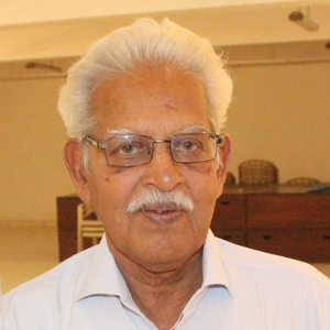An elderly man with white hair and a mustache, wearing glasses and a light-colored collared shirt, stands indoors in a warmly lit room with furniture in the background, reflecting the cozy atmosphere of a home in India.