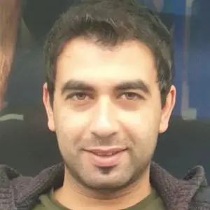 A man with short dark hair and facial stubble is smiling slightly, wearing a dark sweater over a green shirt, and is facing the camera against a blurred background.