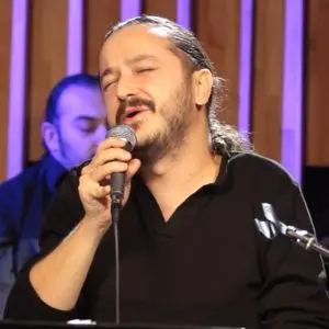 A man with long hair tied back is singing passionately into a microphone. He is wearing a black shirt and appears to be performing indoors, with another musician blurred in the background.