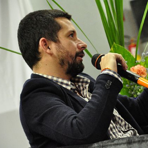 A man with short dark hair and a beard, wearing a dark blazer and checked shirt, holds a microphone while seated indoors with green plants and flowers in the background.