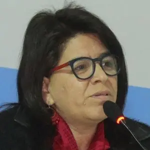 A woman with dark hair and glasses speaks into a microphone. She is wearing a red scarf and dark clothing, and appears to be participating in a discussion or giving a speech.