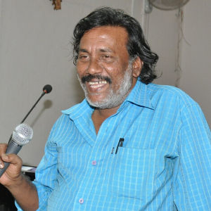 A man with wavy hair and a beard, wearing a blue checked shirt, smiles and speaks into a microphone in an indoor setting. A wall-mounted fan and a microphone stand are visible in the background.