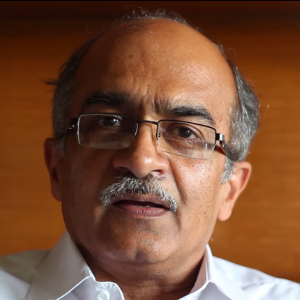 A middle-aged man with glasses, a mustache, and thinning hair is wearing a white shirt and looking directly at the camera, with a blurred orange-brown background.