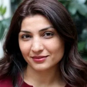 A woman with long, wavy dark brown hair and a slight smile, wearing a dark red top, is pictured in front of a blurred background of green foliage.
