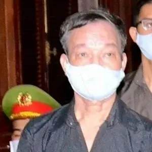 A man wearing a light blue face mask and a black shirt stands indoors, with another masked person and someone in a green hat with a red band in the background.