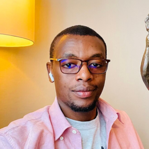 A man wearing glasses, a pink shirt, and wireless earbuds sits indoors near a lamp, looking at the camera with a slight smile.