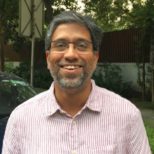 A man with gray hair and glasses, wearing a pink and white striped shirt, smiles while standing outdoors with trees and greenery in the background.