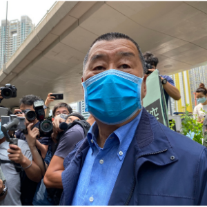 A man wearing a blue face mask and blue jacket stands outdoors while several photographers take pictures of him in an urban setting with tall buildings in the background.