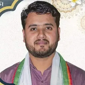 A man with short dark hair and a beard is wearing a purple shirt and a scarf with green, white, and orange stripes, standing in front of a decorative, light-colored background.