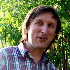 A man with medium-length brown hair wearing a red and white plaid shirt smiles outdoors. Lush green foliage and sunlight are visible in the background.
