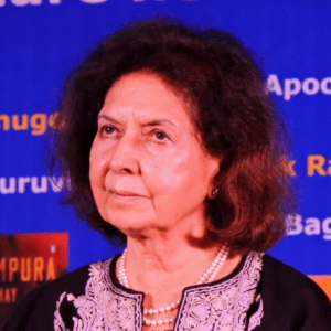 A woman with wavy dark hair wearing a black outfit with white embroidery and a pearl necklace stands in front of a blue background with partially visible yellow and white text.