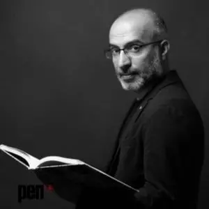 Black and white photo of a man with glasses and a beard, wearing a dark jacket and holding an open book, looking over his shoulder toward the camera. The PEN logo is visible in the lower left corner.