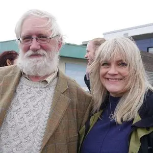 An older man with white hair, a beard, glasses, and a patterned sweater stands beside a smiling woman with long blonde hair and a jacket. They are outdoors, with other people and buildings in the background.
