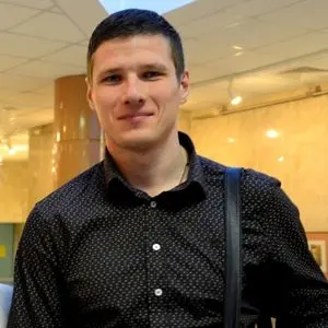 A man with short dark hair wearing a black, patterned button-up shirt and a shoulder bag strap stands indoors, smiling slightly. The background features warm lighting and a tan, modern interior.